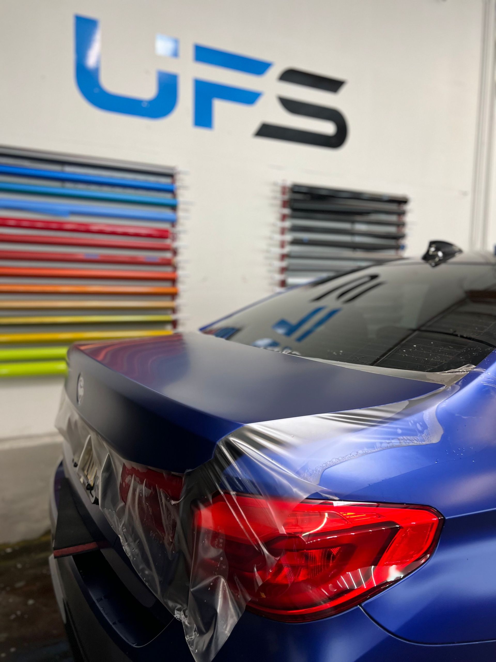 Blue sports car in a workshop, rear view, with colorful vinyl rolls on the wall and UFS sign nearby