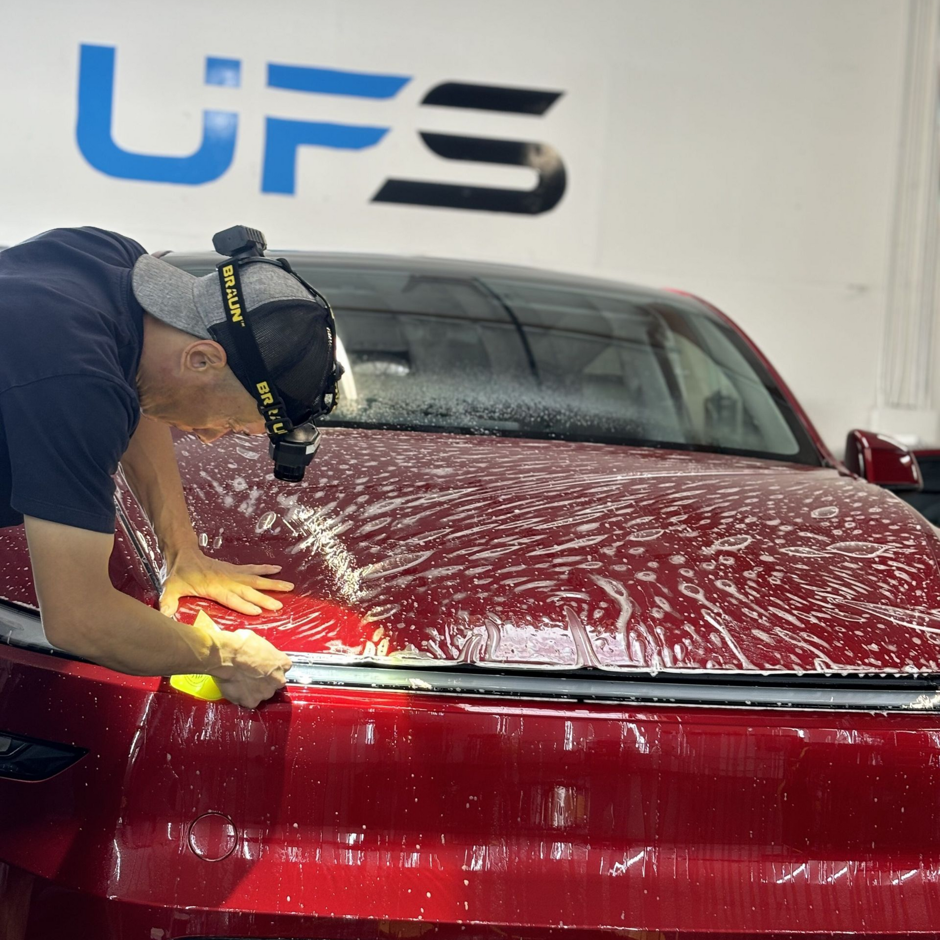 Technician polishing a red car hood in a detailing shop under a UFS sign