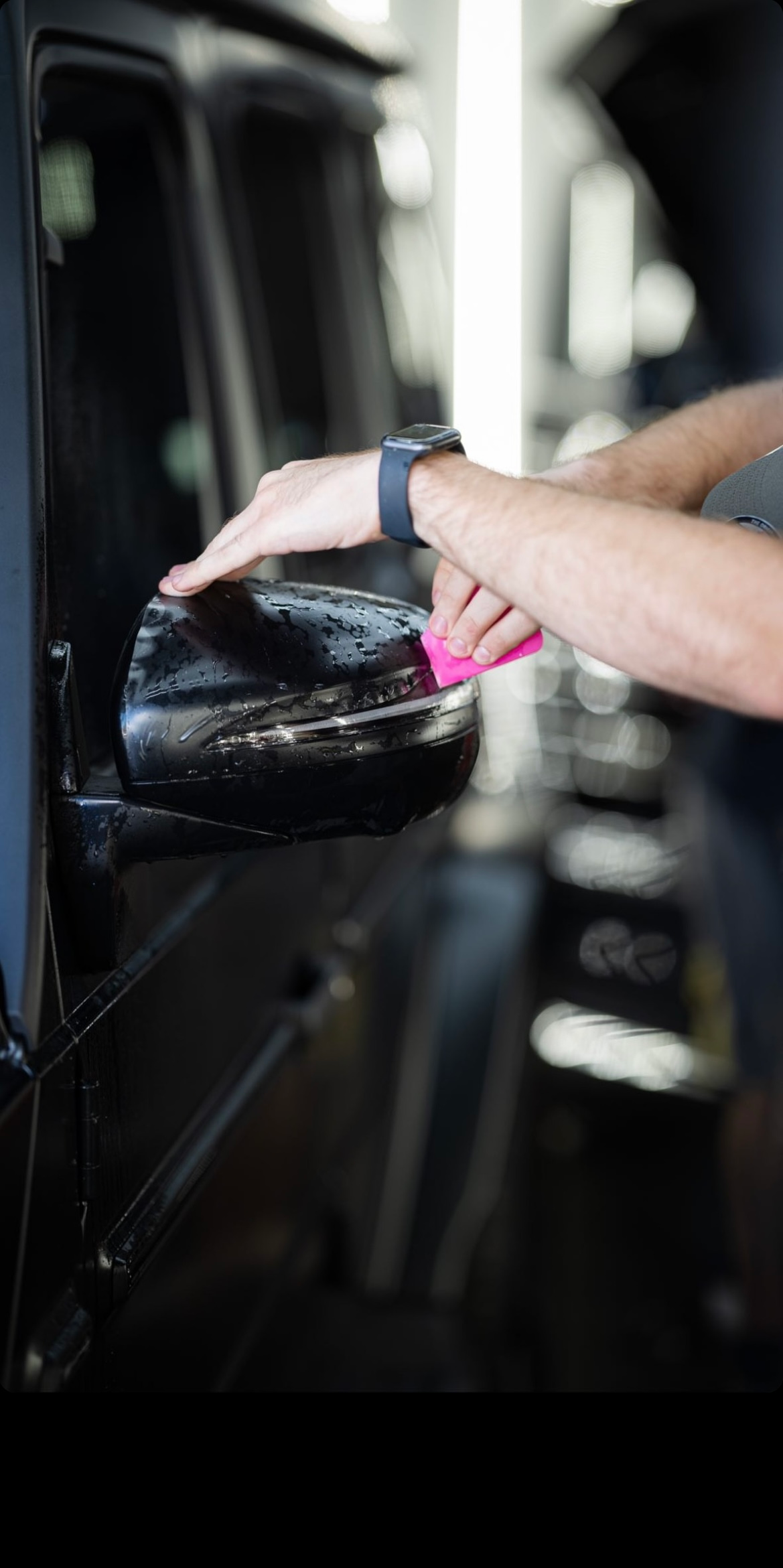 Person hand-washing a black car door with a pink cloth in a garage