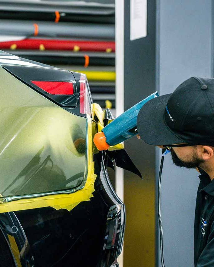 Technician polishing a black car’s rear quarter panel with a blue buffer in a workshop
