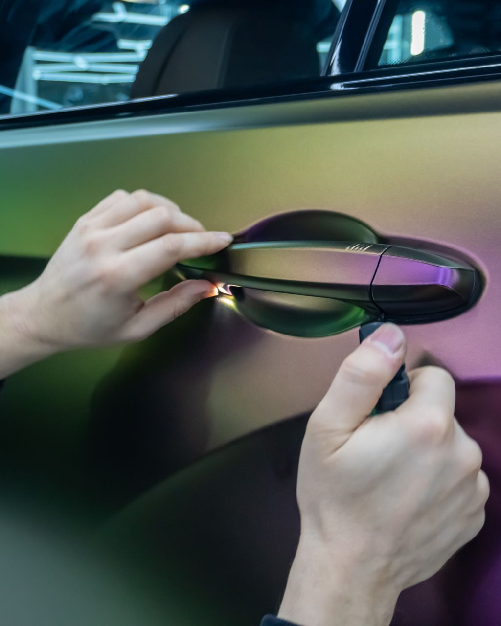 Hands peeling protective film from a car door handle on a metallic surface.