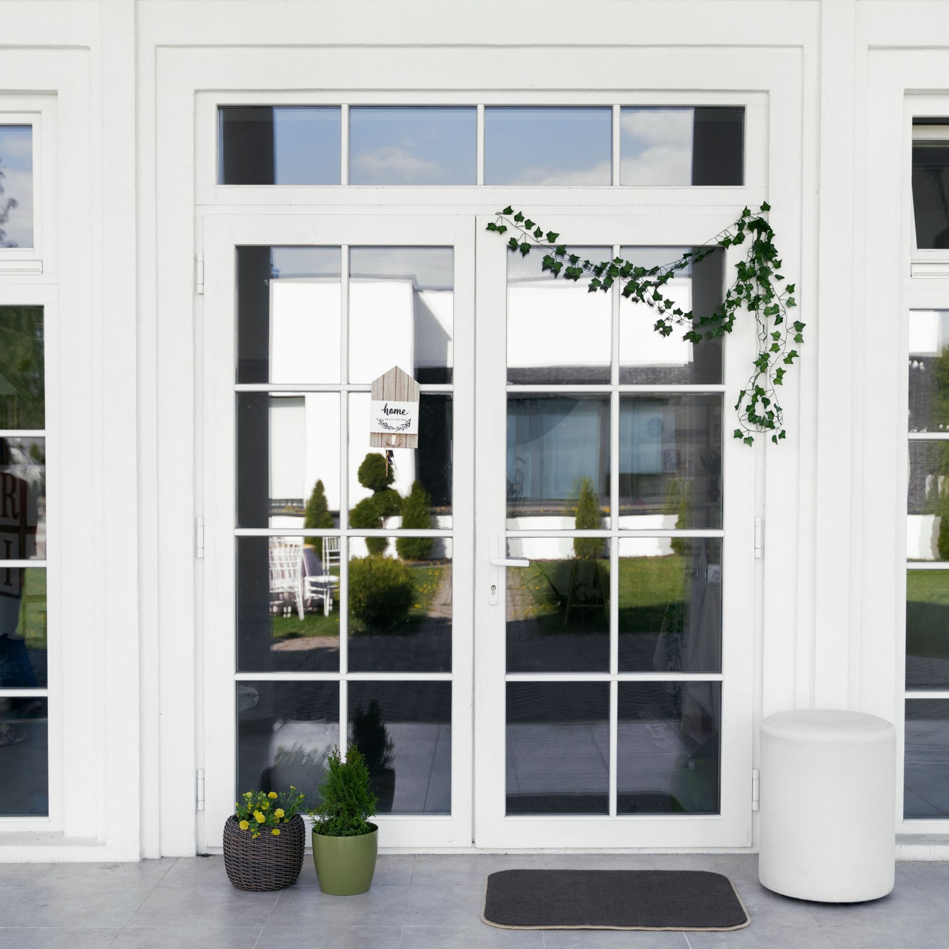 White storefront entrance with glass double doors, hanging greenery, and potted plants by the doorway