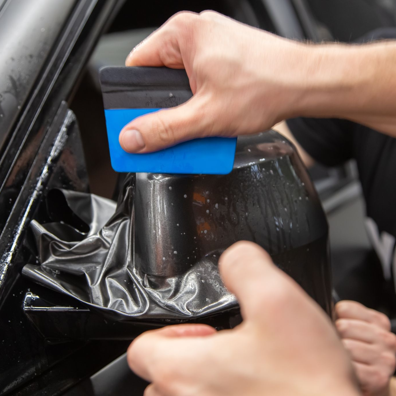 Hands using a blue squeegee to smooth black vinyl film on a car window