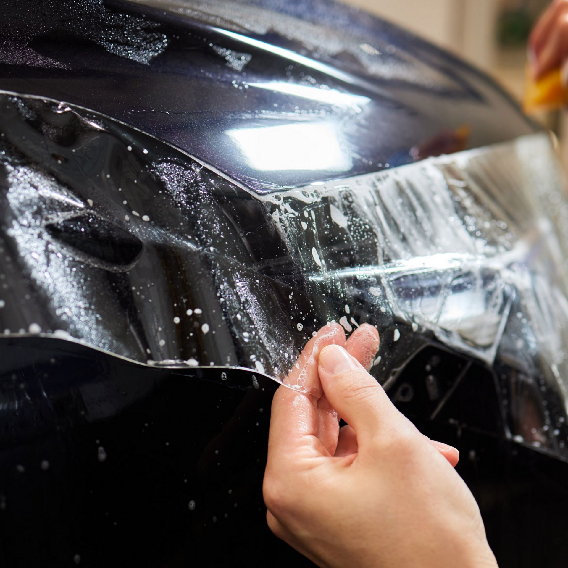 Person peeling protective film from a glossy black car hood during detailing