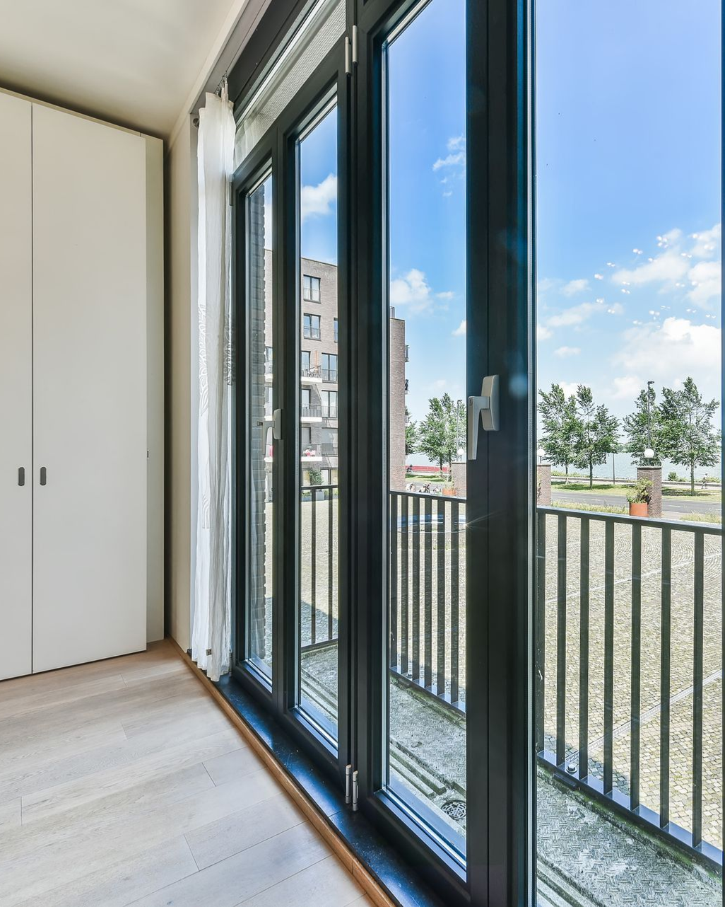 Bright room with large black-framed windows opening to a balcony and trees outside