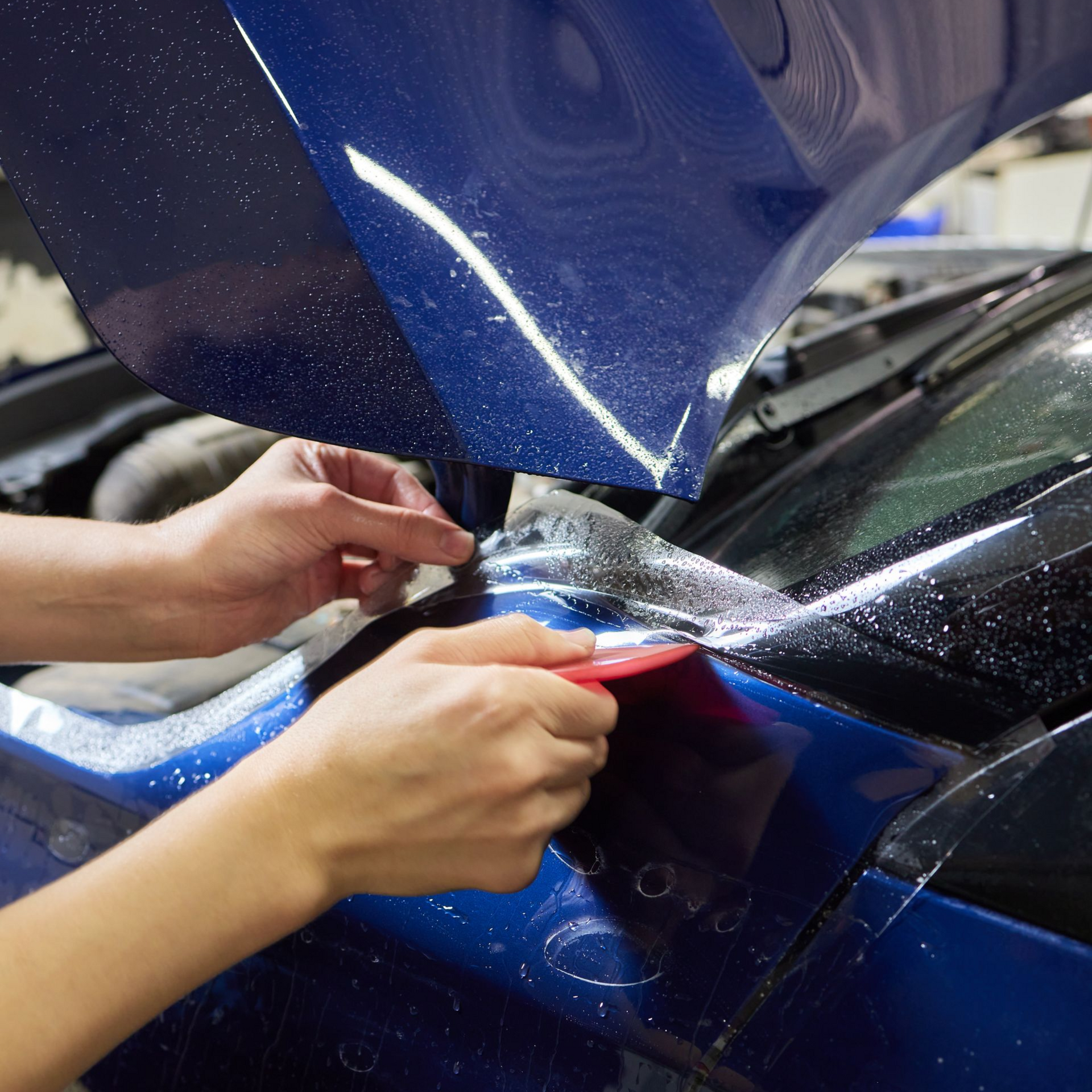 Hands applying red tape to a blue car panel in a workshop