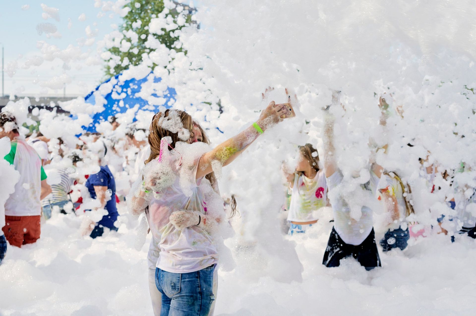 People at a foam party; woman reaches up, surrounded by white foam. Outdoors, sunny.