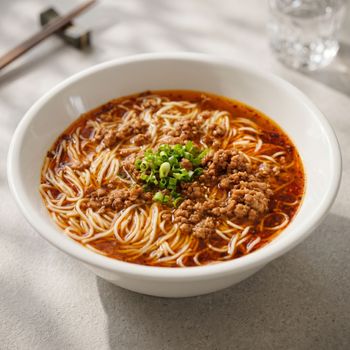 Bowl of noodles in red broth topped with minced meat and green onions on a table