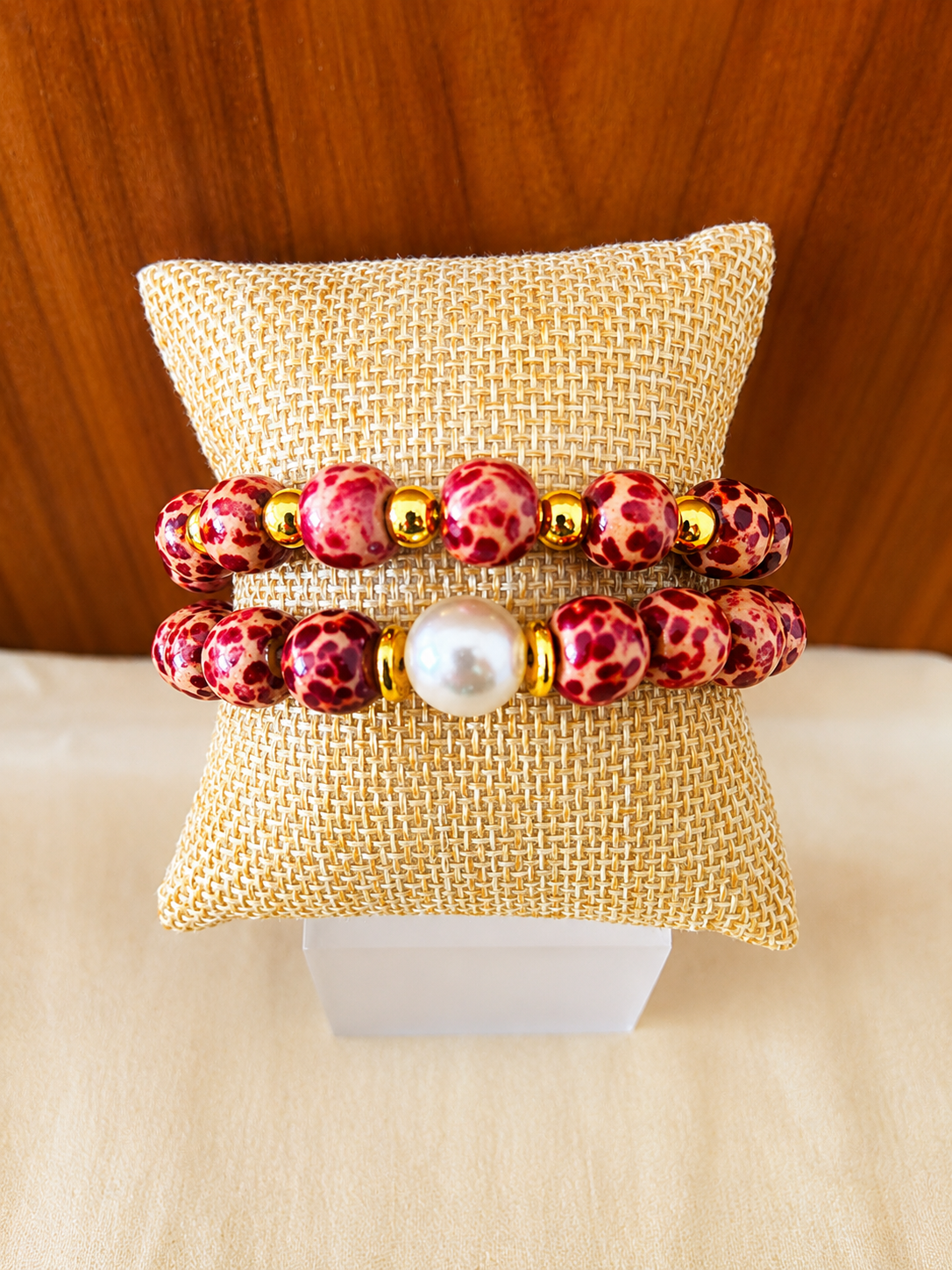 Two beaded bracelets, red and pink with gold accents, displayed on a small cushion