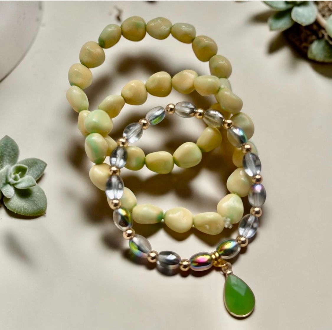 Three beaded bracelets in cream, green, and iridescent tones, one with a green teardrop charm.