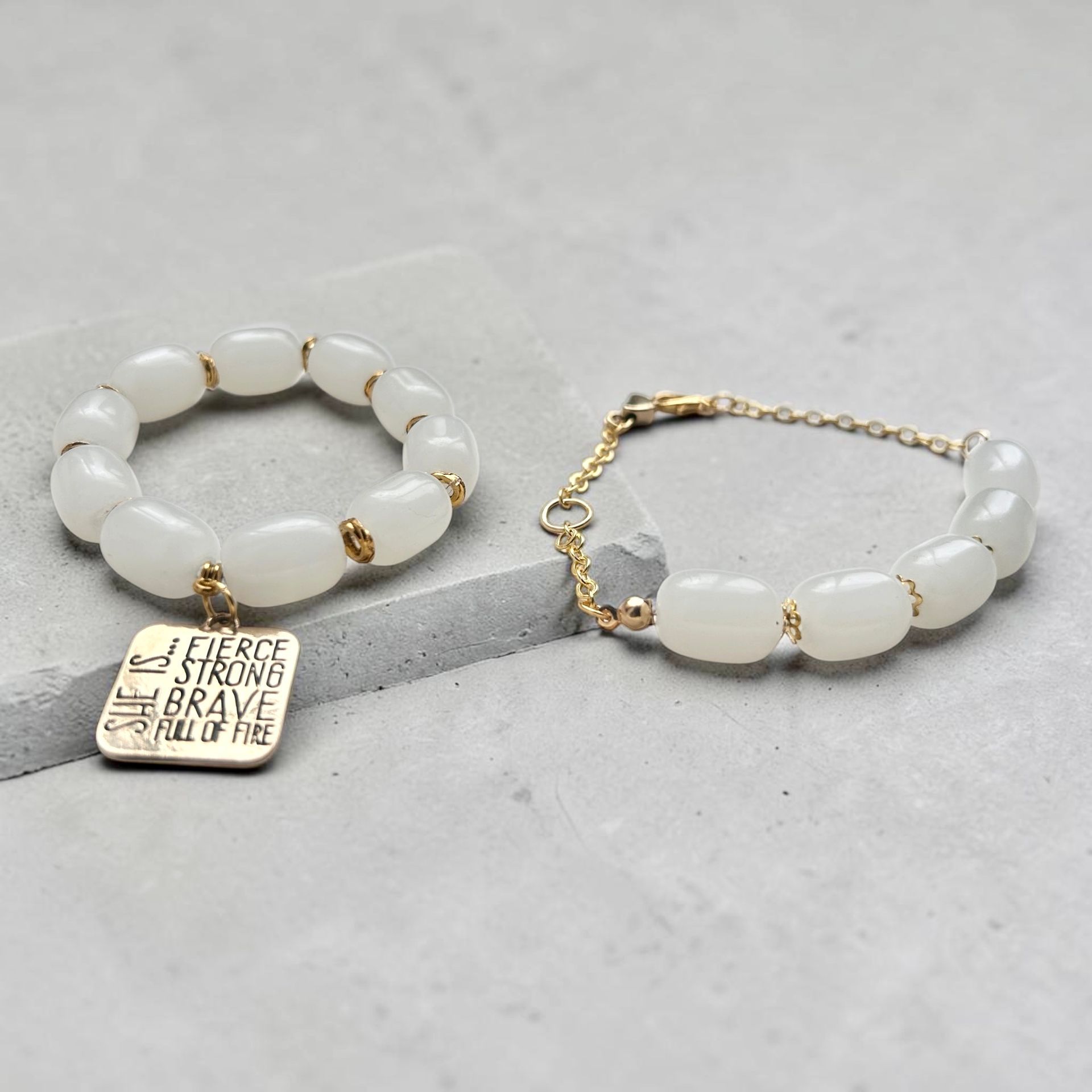 Two white beaded bracelets, one with a gold engraved charm, on a light surface