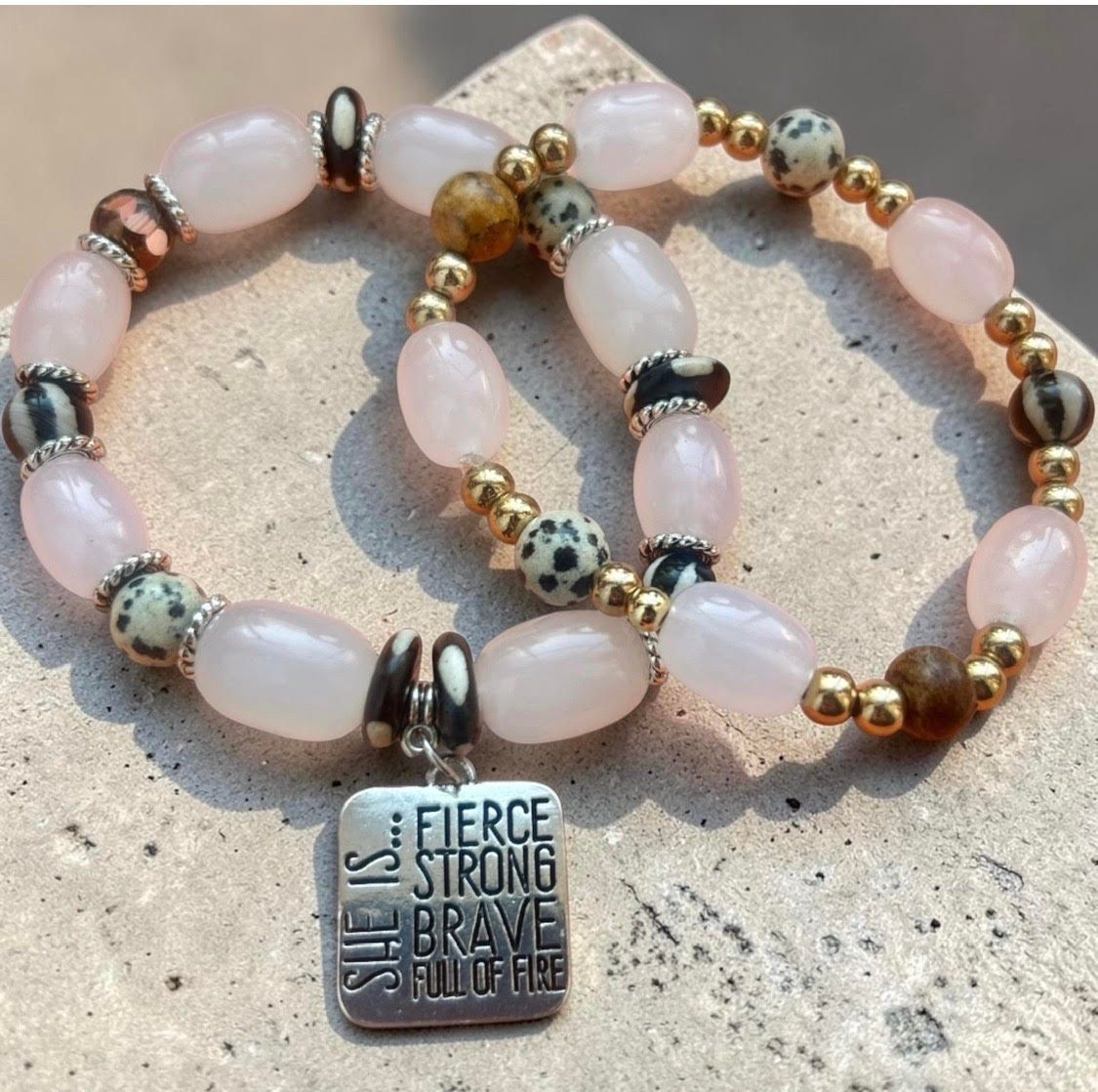 Three beaded bracelets with pink stones and a silver charm reading “Fierce, Strong, Brave” on a stone surface