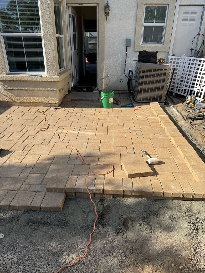A partially installed tan brick patio sits in front of a house entrance, with an orange power cord and a green bucket.
