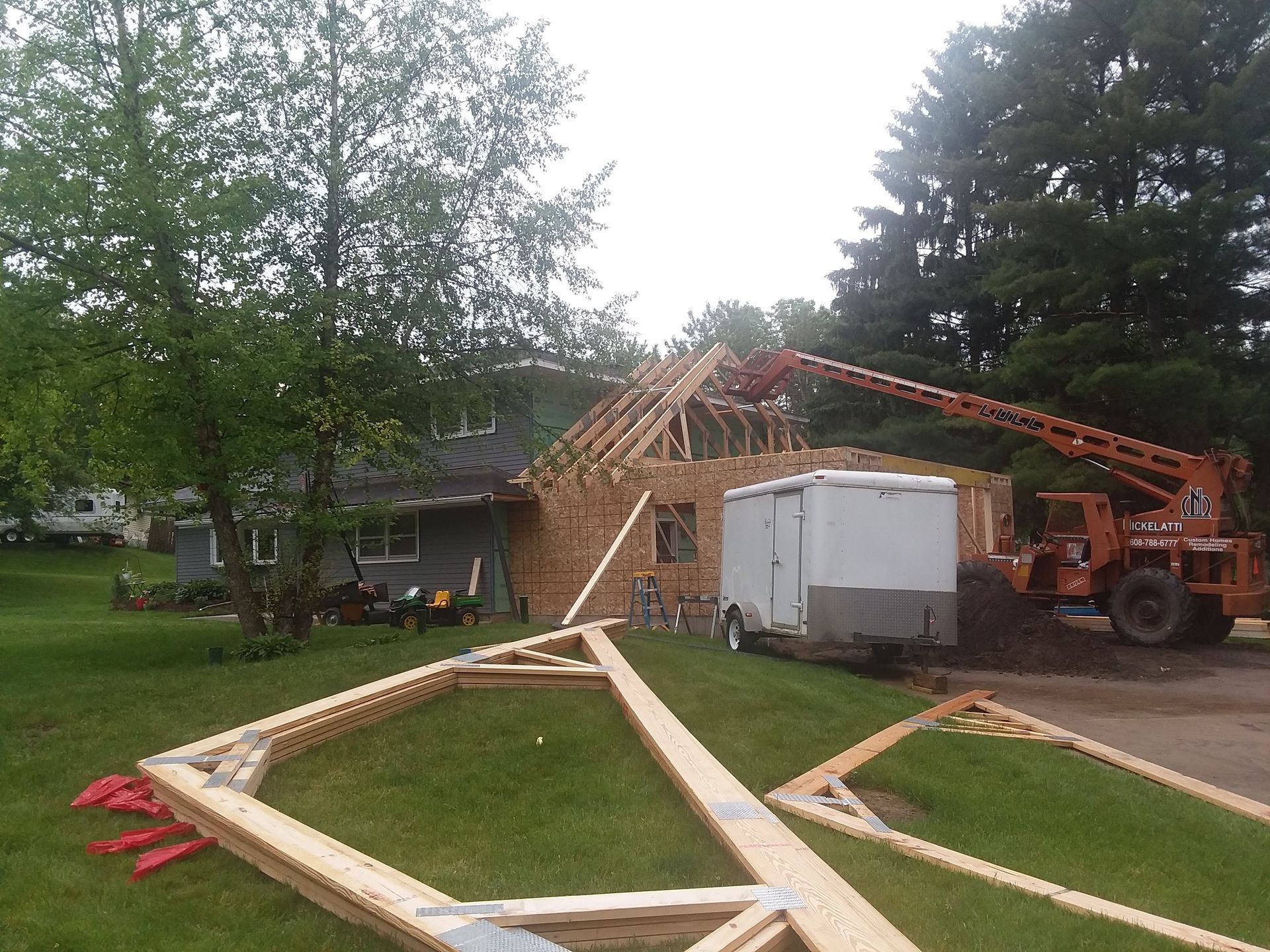 Roof framing under construction with crane lifting trusses near a house and trailer in a yard