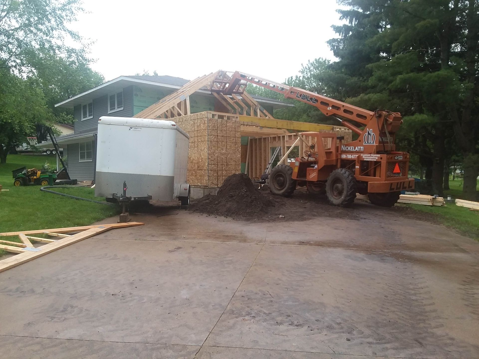 Orange forklift lifting roof trusses beside a house under construction, with a white trailer nearby.
