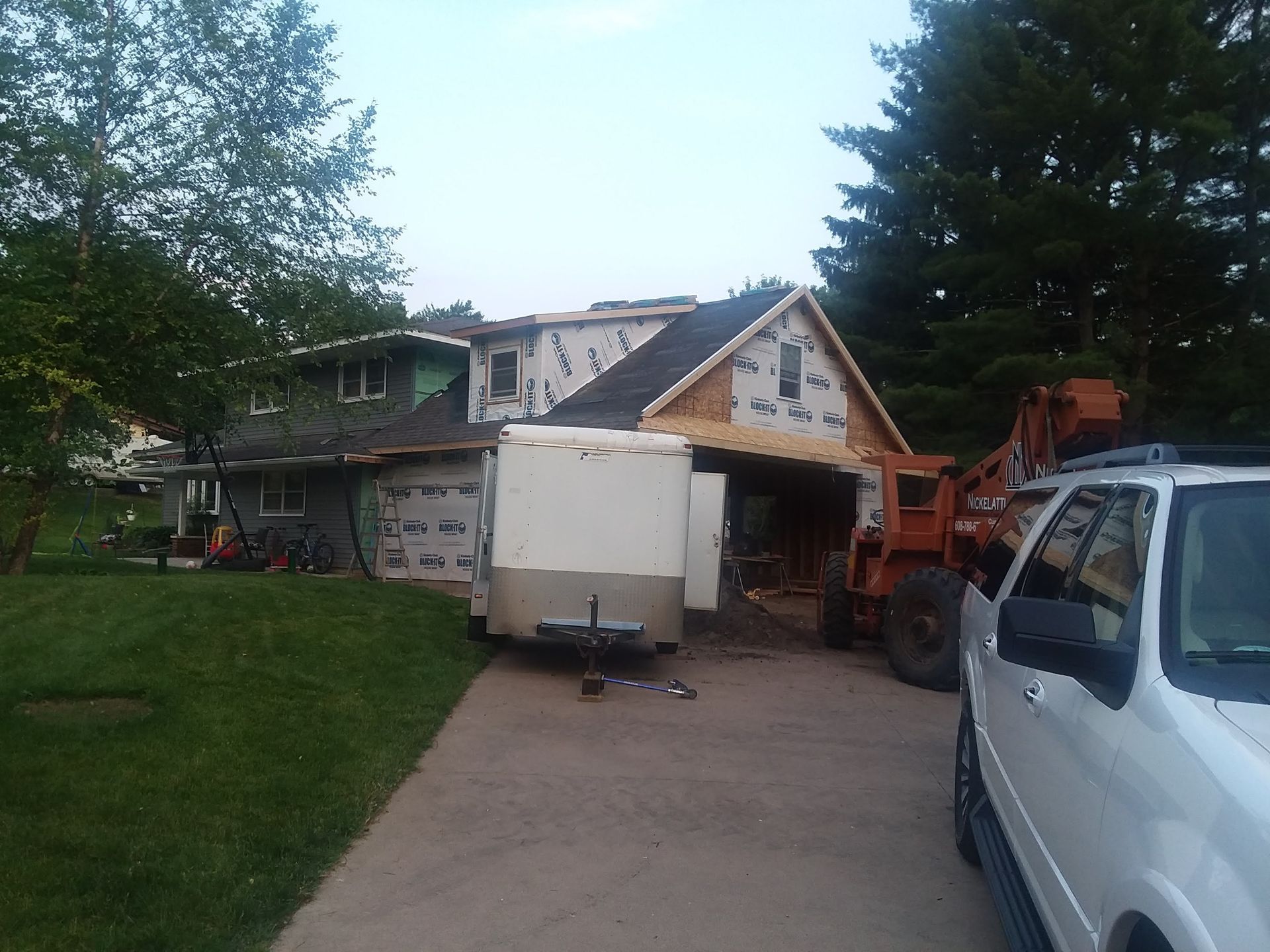 House under construction with a trailer and orange excavator in a driveway, white vehicle at right