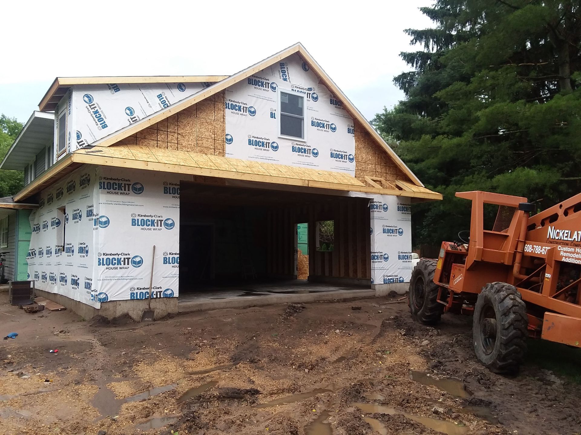 Partially built house wrapped in white house wrap, with muddy ground and an orange tractor at right