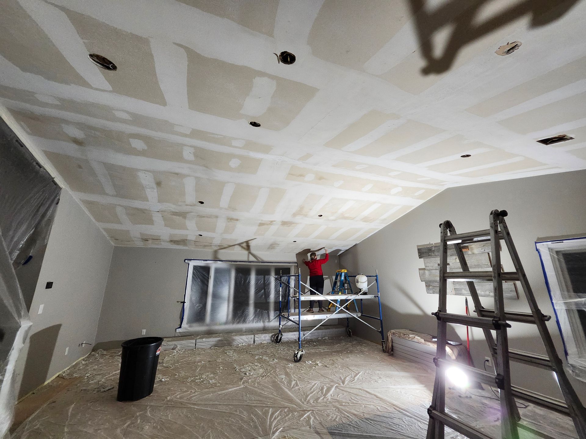 Room under renovation with taped ceiling, ladders, and workers near a window while flooring is covered.