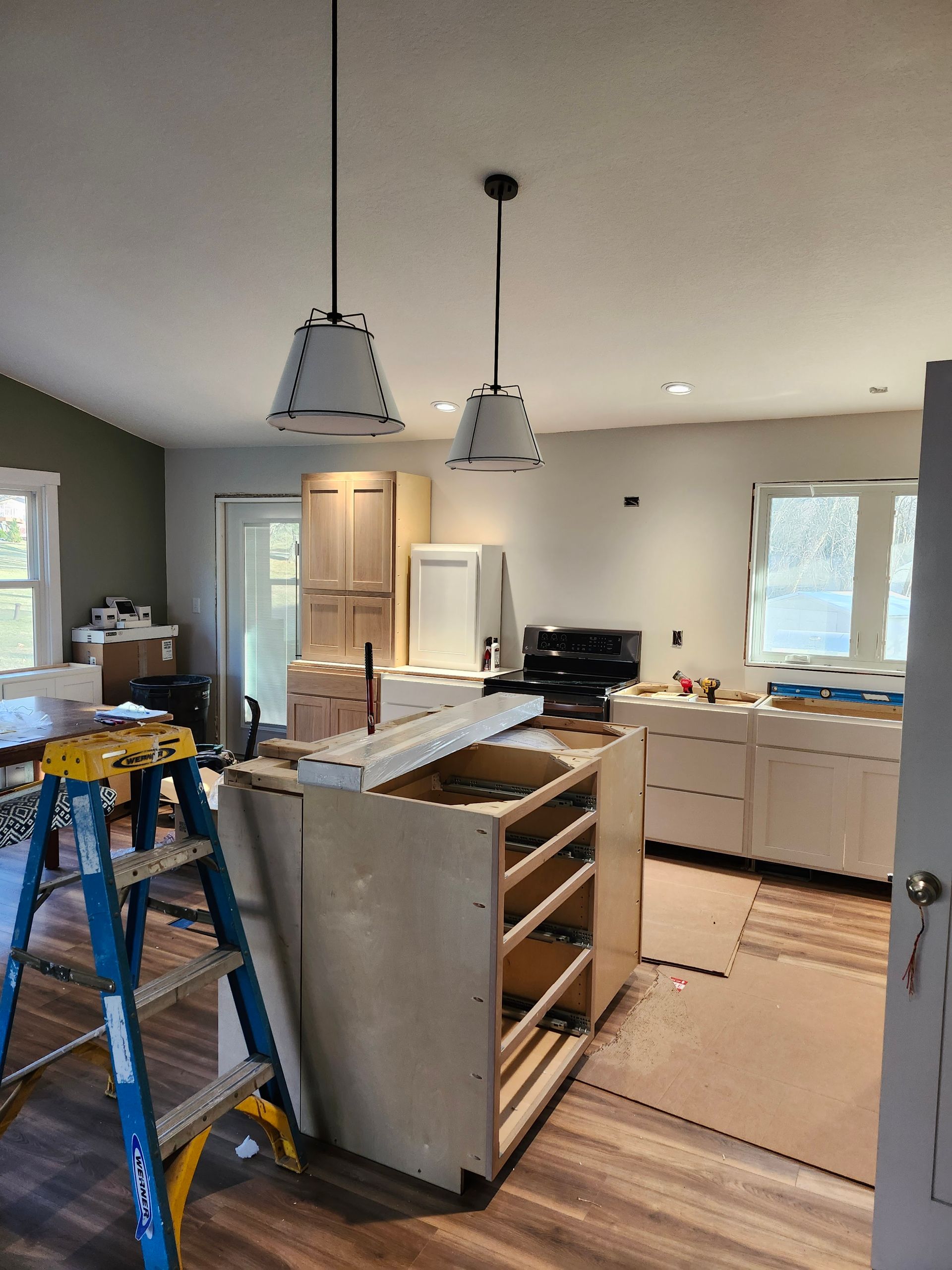 Kitchen under renovation with open cabinets, island, ladders, and hanging pendant lights