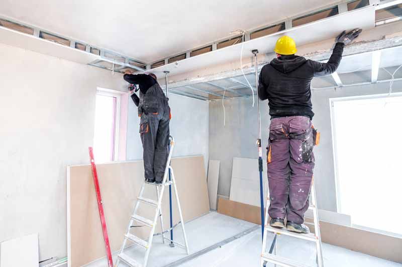 Two workers on ladders installing ceiling drywall in a bright room under renovation