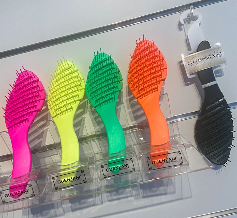 Cinque spazzole per capelli colorate esposte su uno scaffale: rosa, giallo, verde, arancione e nero, con un supporto di plastica trasparente.