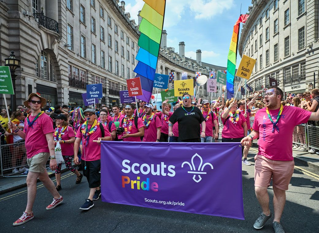 Scouts members marching proudly at London Pride. Scouts Pride group marching at London Pride, wearing pink shirts and rainbow accessories, carrying a large purple Scouts Pride banner and rainbow flags through a busy city street.