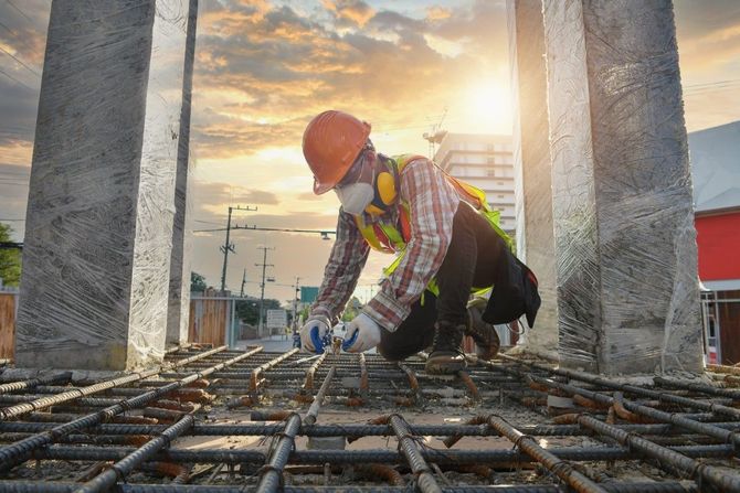 Construction worker in orange hard hat crouches on rebar at a building site during sunset