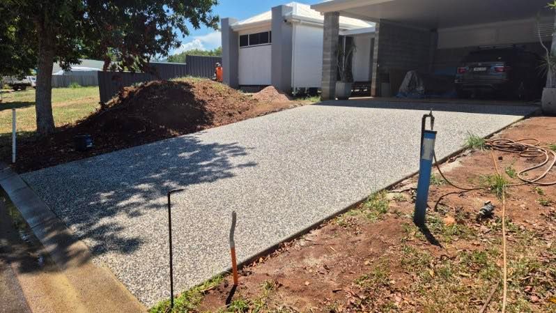 Concrete driveway leading to a covered carport beside a white house, with dirt landscaping and a tree. — King Reef Concrete in Kurrimine Beach, QLD