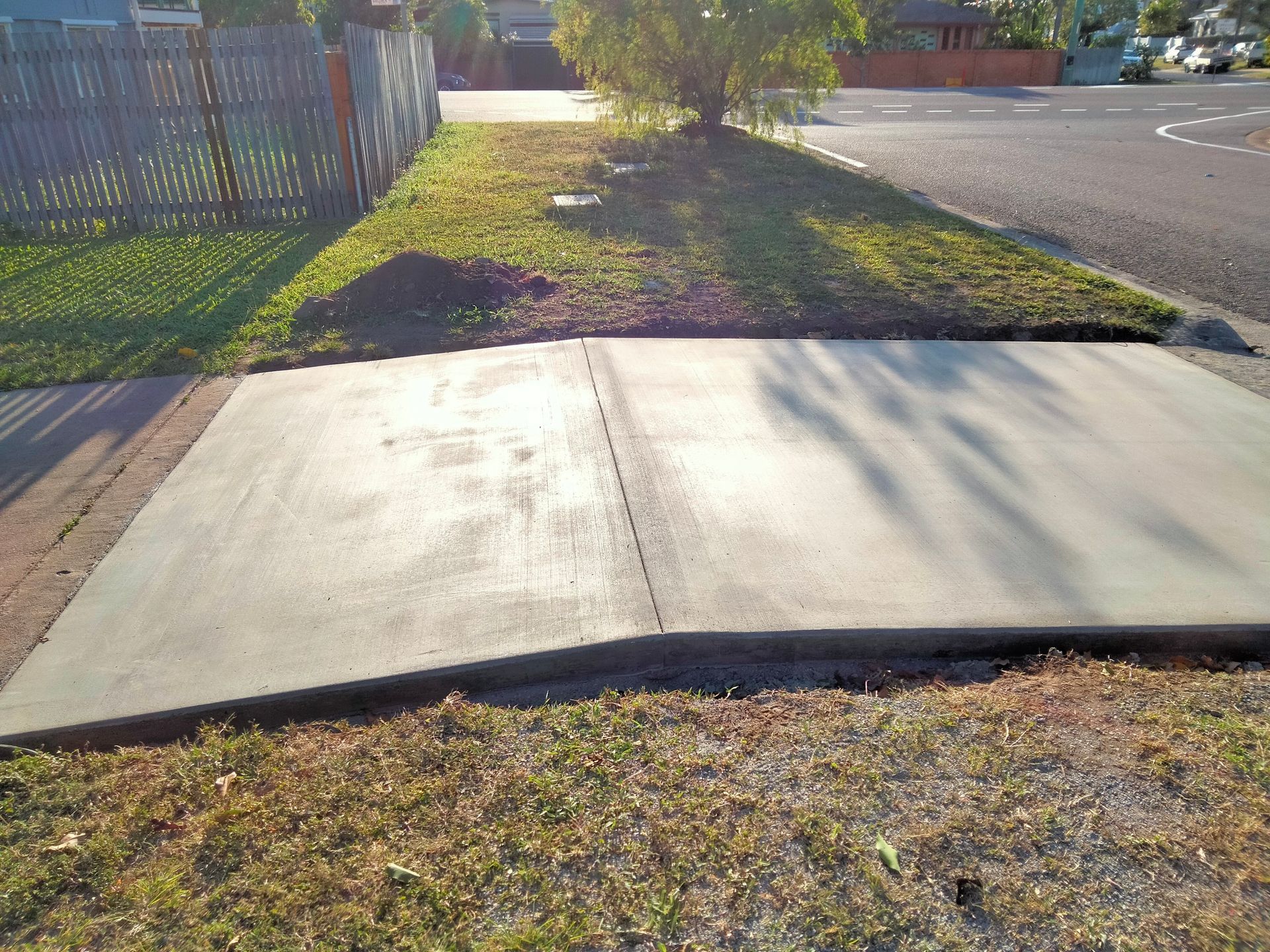 Broken sidewalk ramp at a curb, with cracked concrete slabs beside a chain-link fence and grassy verge. — King Reef Concrete in Kurrimine Beach, QLD