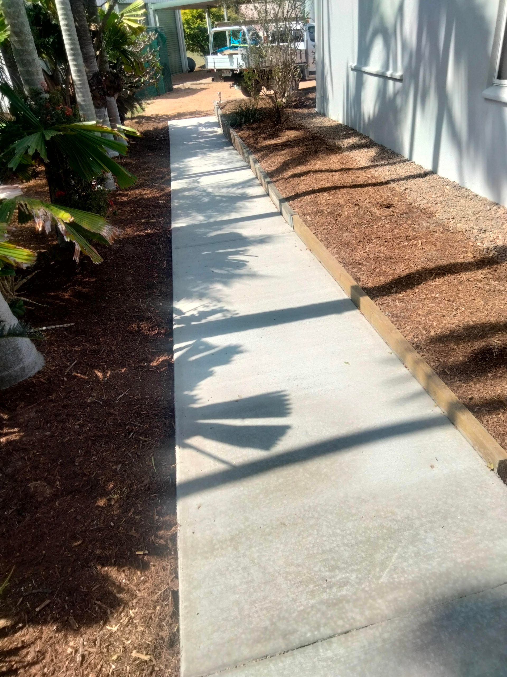 Narrow concrete walkway beside a building, lined with mulch, shrubs, and palm trees in bright sunlight — King Reef Concrete in Kurrimine Beach, QLD