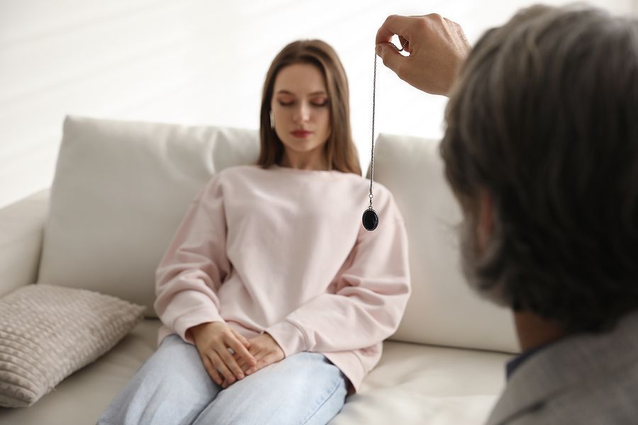 A practitioner holding a swinging pendant in front of a person sitting on a white sofa during a hypnotherapy session.