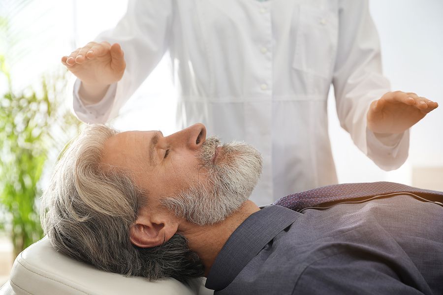 A person in a white lab coat holds their hands over a reclining person's head during a therapeutic energy session.