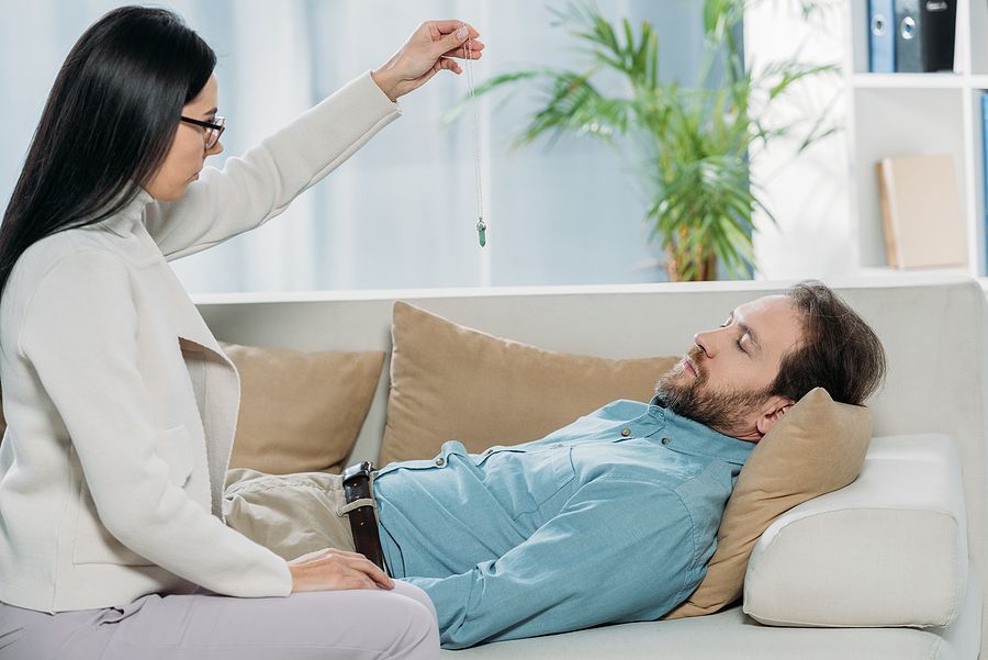 A person in a light-colored sweater holds a pendulum above a person resting on a sofa during a hypnotherapy session.
