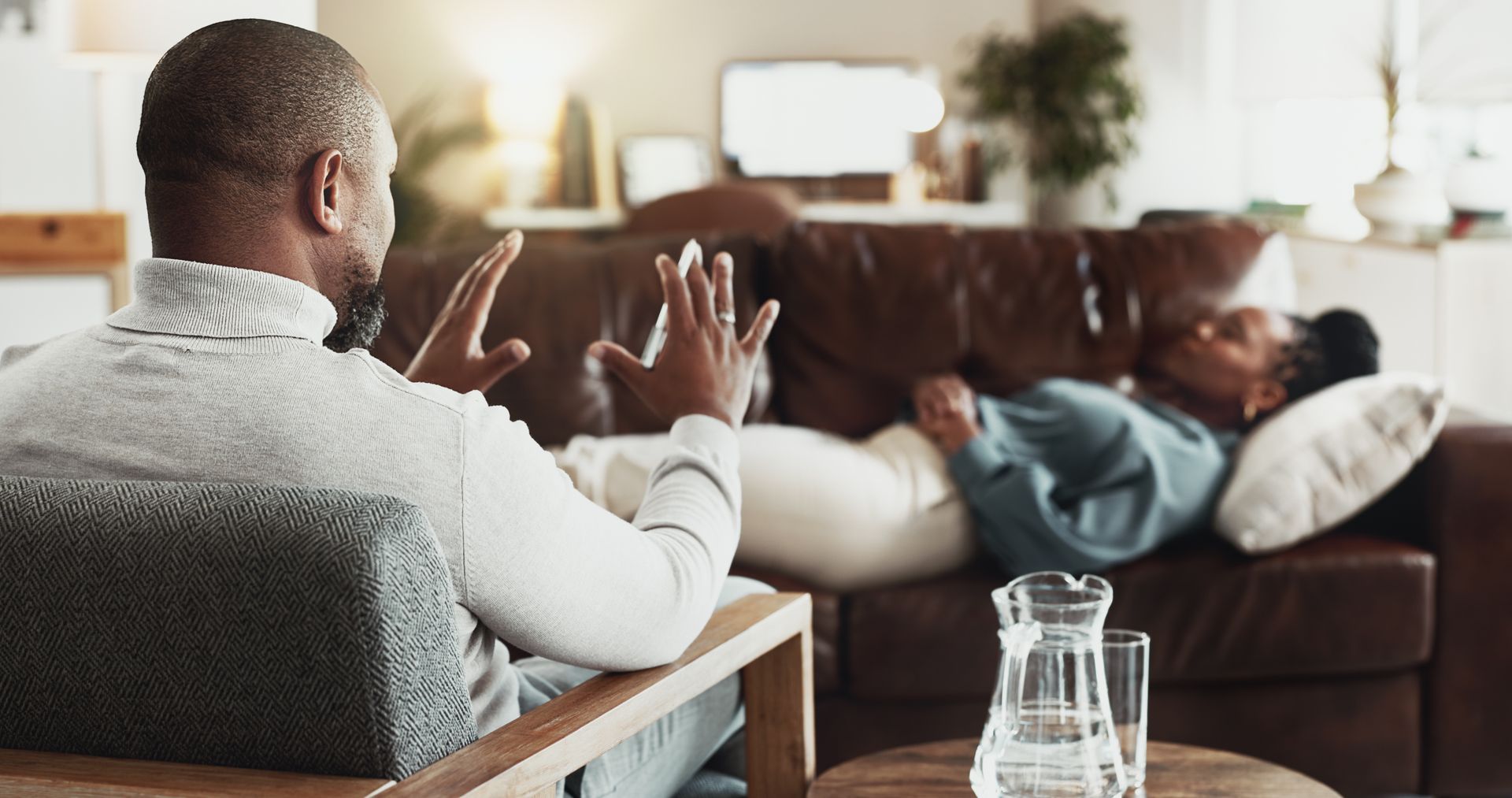 A person sits in a chair gesturing while a patient lies on a couch during a therapy session in a living room.