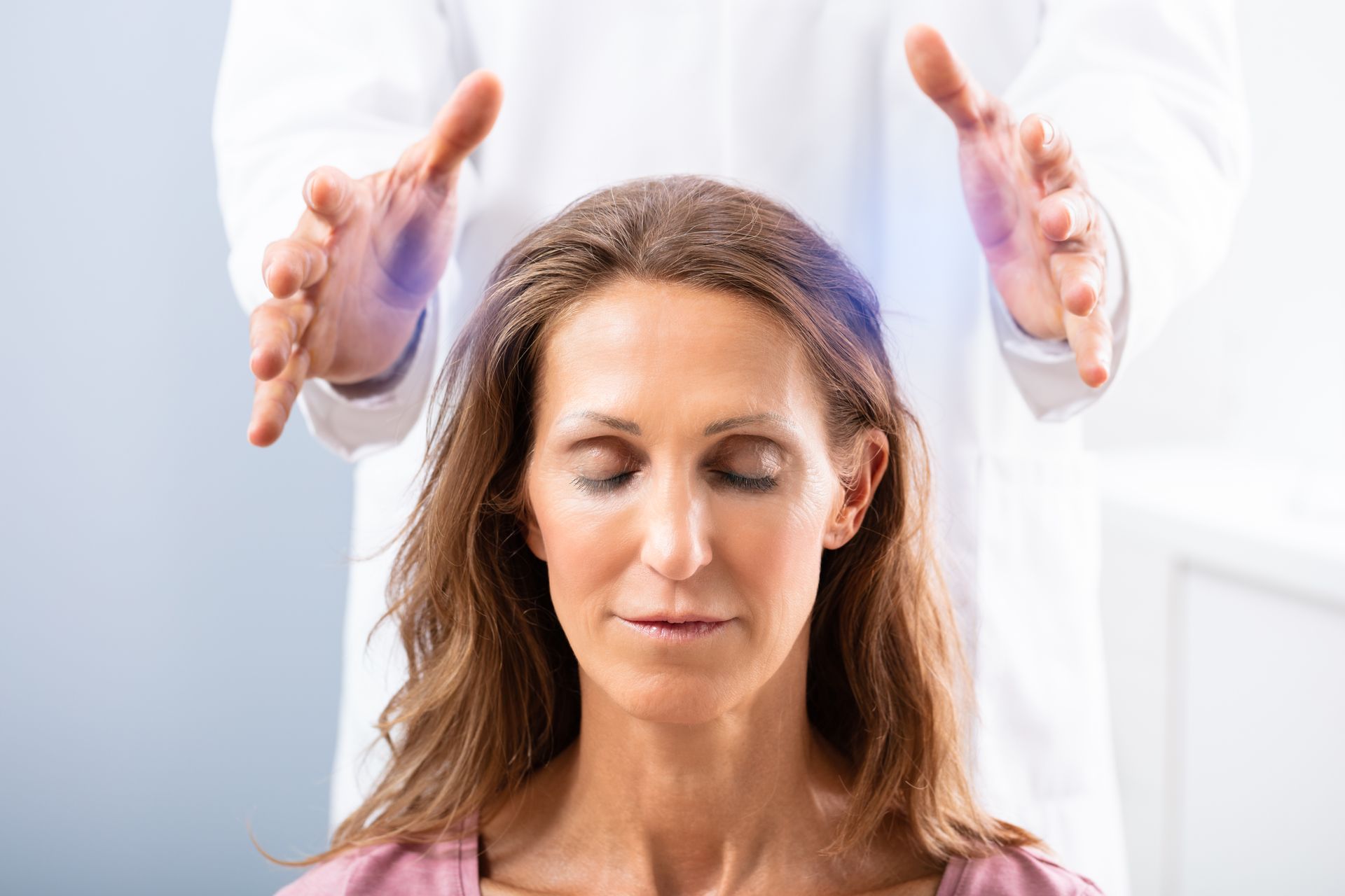 A practitioner in a white coat holds hands above a person’s head, who sits with eyes closed in a calm, focused state.