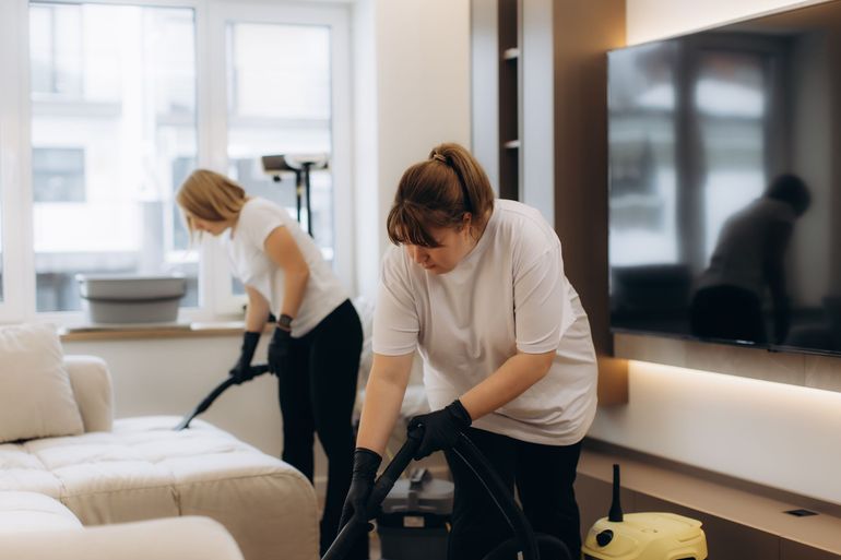 Two cleaners vacuuming a bright living room with a large TV and white sofa.