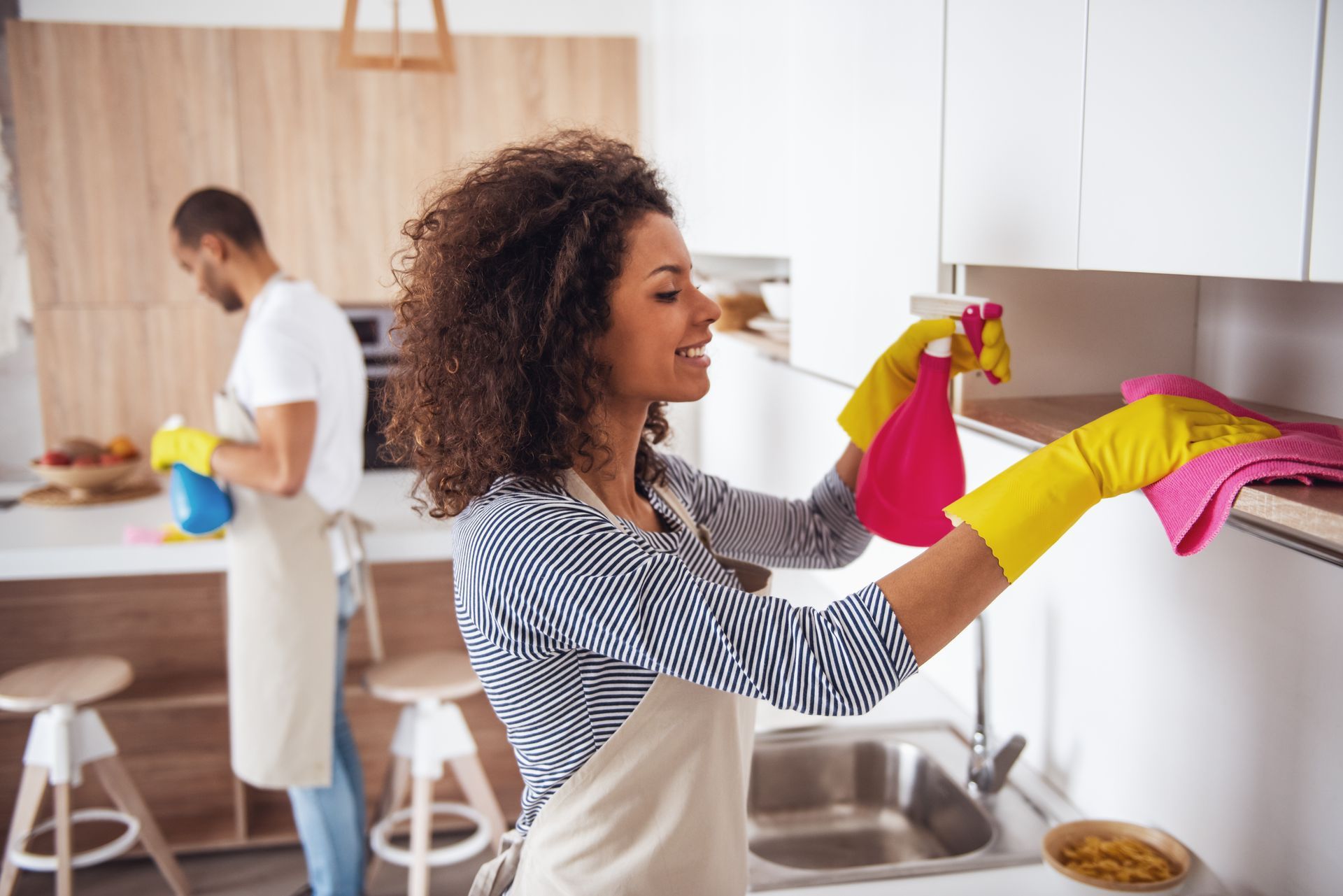 Woman cleaning kitchen cabinets with spray bottle and cloth while another person washes dishes in the background