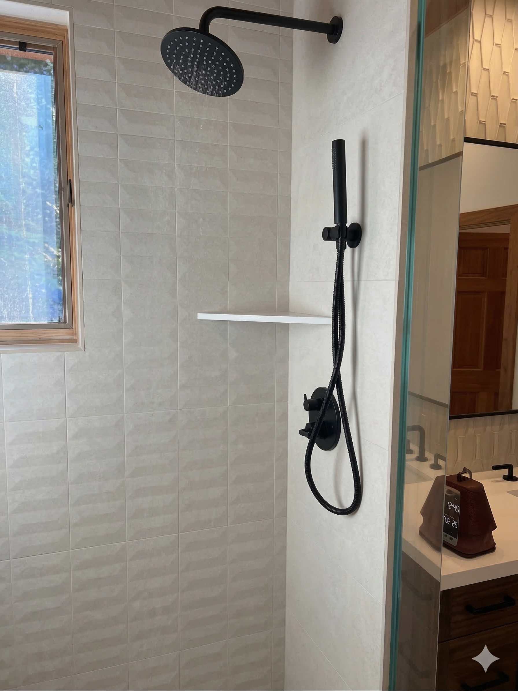 A matte black rainfall shower head and handheld wand fixture mounted on a wall with white textured tiles and a shelf.