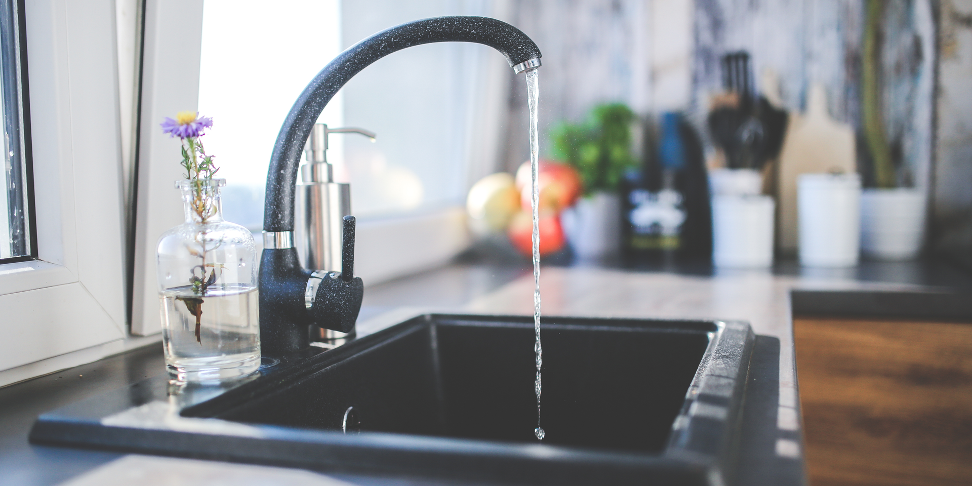 Water flows from a black kitchen faucet into a dark sink next to a vase with a small flower on a window sill.