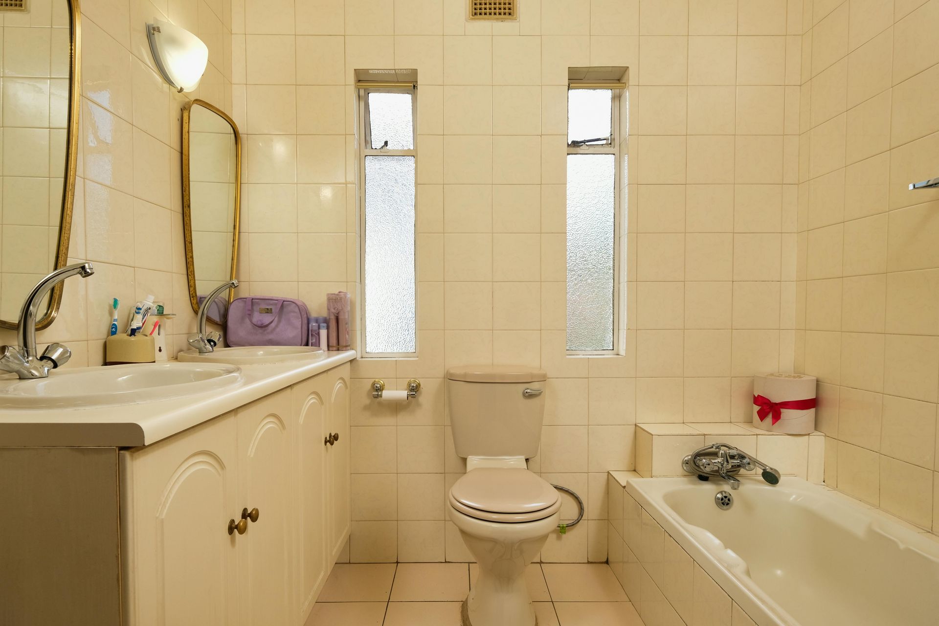 A beige tiled bathroom with a double vanity, toilet, bathtub, and two narrow windows.