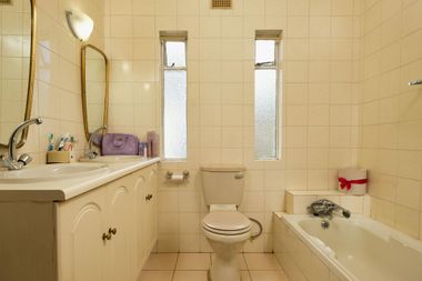 A beige tiled bathroom with a double vanity, toilet, bathtub, and two narrow windows.