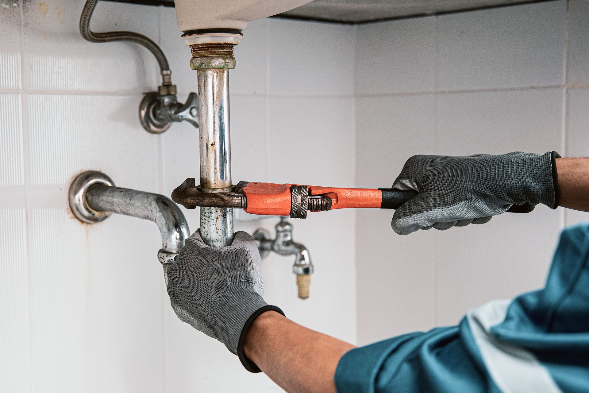 A plumber wearing work gloves uses an orange pipe wrench to tighten metal plumbing pipes under a white-tiled sink.