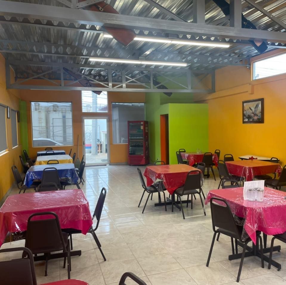 A dining area with tables covered in red, pink, and yellow cloths, metal chairs, tile floors, and high industrial ceilings.