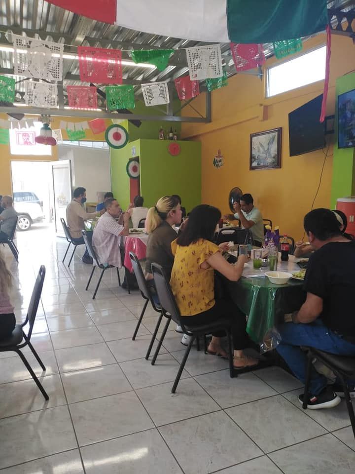 People sit at tables eating and chatting in a small restaurant decorated with Mexican flag-colored banners.