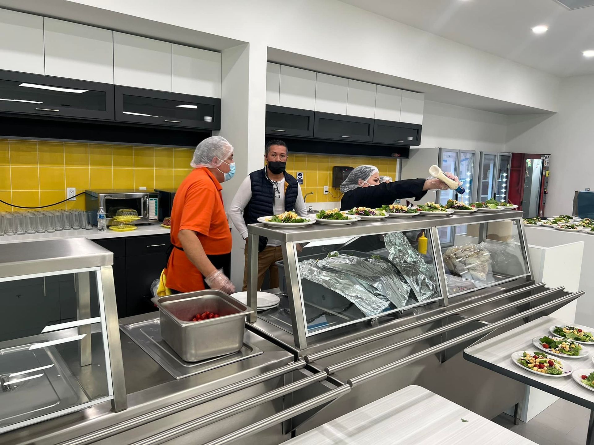 Three food service workers prepare plates of food behind a stainless steel counter in a kitchen with yellow tiling.