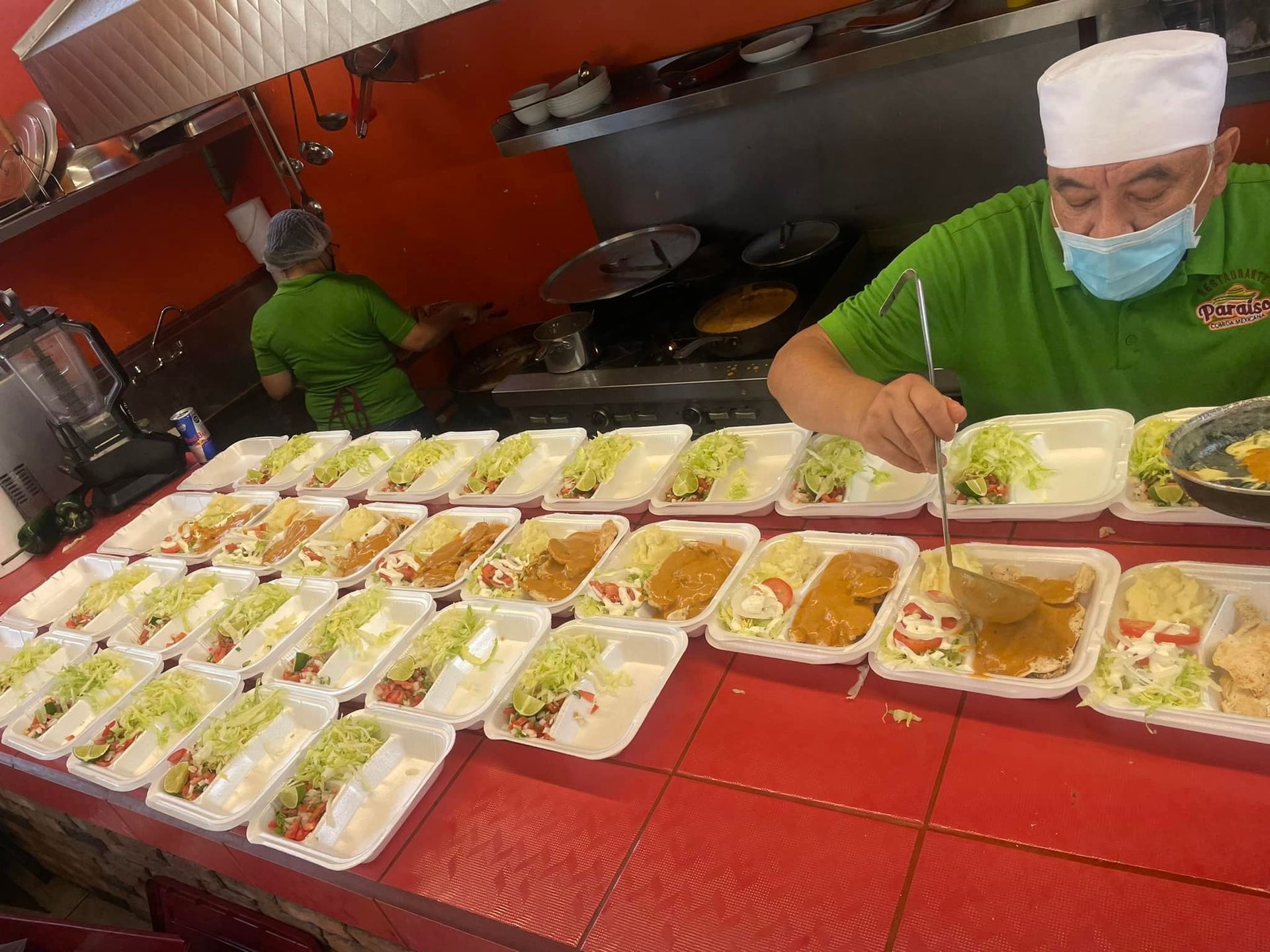 A person wearing a green shirt and face mask dishes food into multiple take-out containers lined up on a red counter.