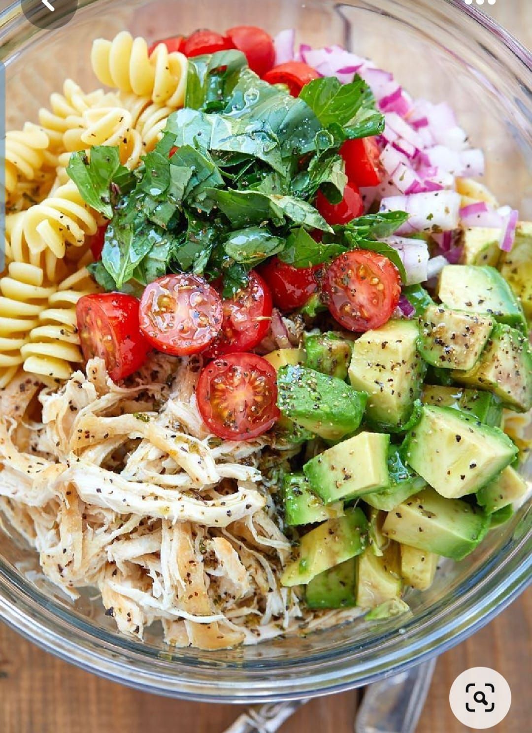 A glass bowl filled with rotini pasta, shredded chicken, chopped avocado, cherry tomatoes, red onion, and fresh herbs.