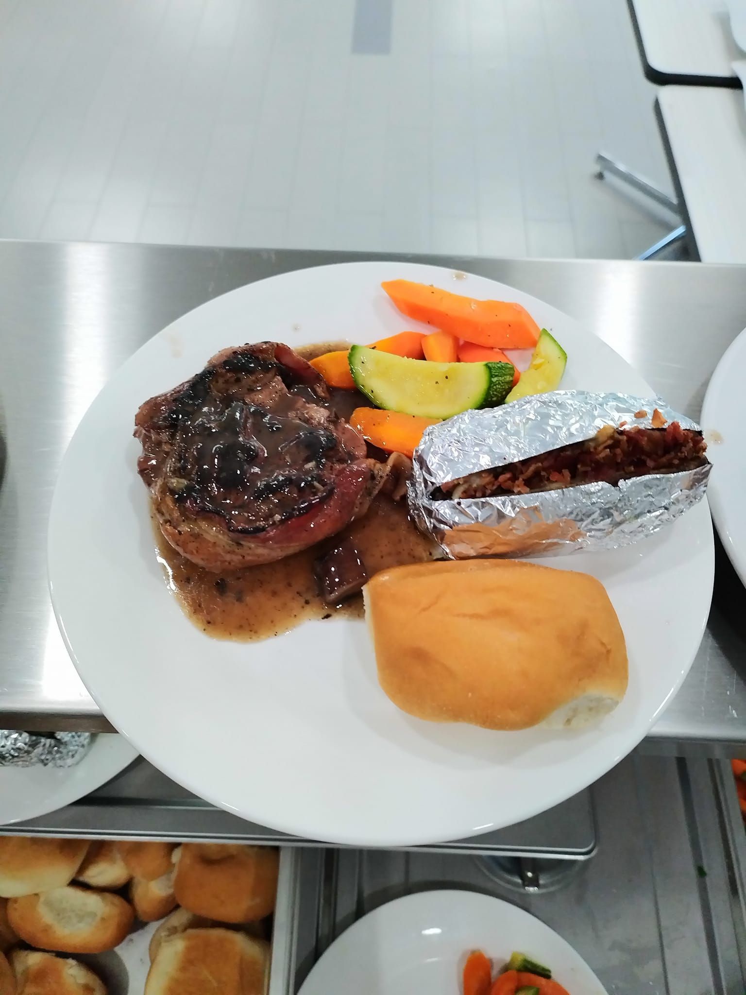 A dinner plate with a meat portion in sauce, a foil-wrapped baked potato, steamed vegetables, and a dinner roll.