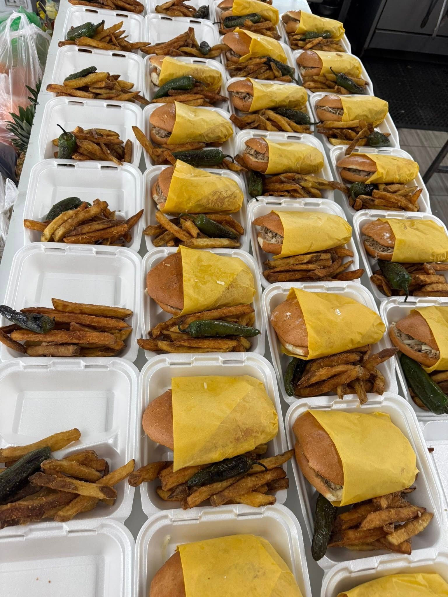 Many styrofoam takeout containers are filled with cheeseburgers wrapped in yellow paper and a side of fries.
