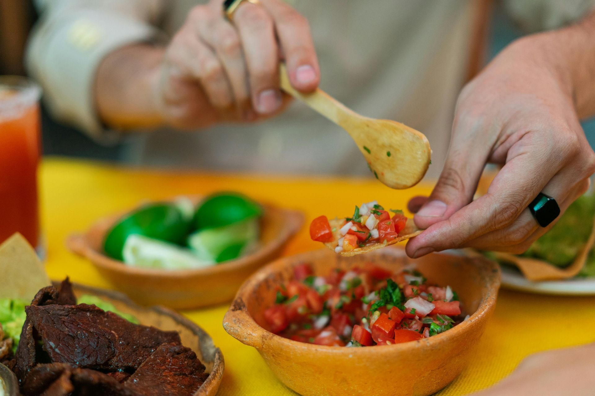 Hands holding a chip scoop pico de gallo from a clay bowl, with lime wedges and meat nearby on a yellow table.